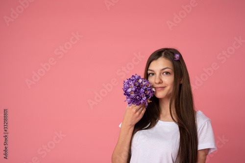 Cheerful brunette female in white t-shirt holding spring snowdrops flowers and looking at the camera, isolated over pink background