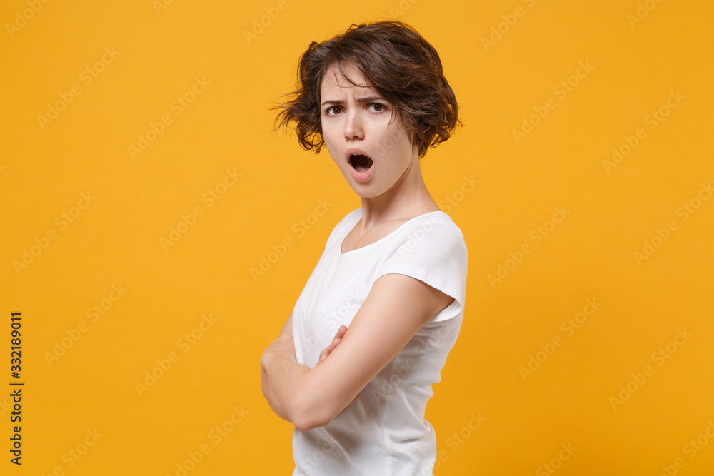 Side view of shocked young brunette woman in white t-shirt posing isolated on yellow orange background in studio. People lifestyle concept. Mock up copy space. Holding hands crossed, looking camera. Side view of shocked young brunette woman in white t-shirt posing isolated on yellow orange background in studio. People lifestyle concept. Mock up copy space. Holding hands crossed, looking camera.