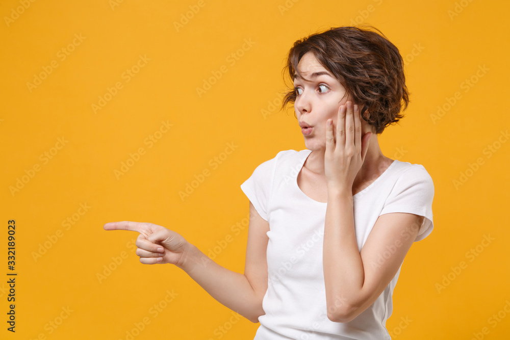 Shocked young brunette woman in white t-shirt posing isolated on yellow orange background studio portrait. People lifestyle concept. Mock up copy space. Pointing index finger aside, put hand on cheek. Shocked young brunette woman in white t-shirt posing isolated on yellow orange background studio portrait. People lifestyle concept. Mock up copy space. Pointing index finger aside, put hand on cheek.