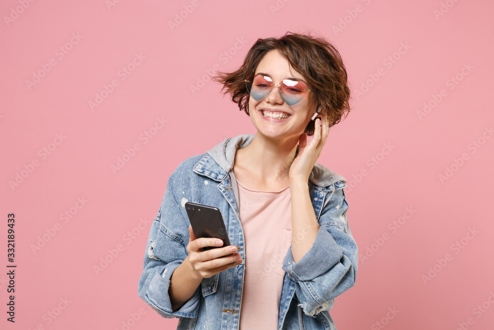 Smiling young brunette woman in casual denim jacket, eyeglasses posing isolated on pastel pink background. People lifestyle concept. Mock up copy space. Listen music with air pods, hold mobile phone. Smiling young brunette woman in casual denim jacket, eyeglasses posing isolated on pastel pink background. People lifestyle concept. Mock up copy space. Listen music with air pods, hold mobile phone.