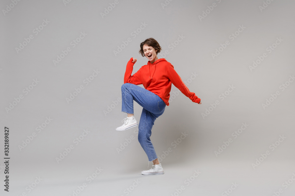 Side view of happy young brunette woman girl in casual red hoodie blue jeans posing isolated on grey background. People lifestyle concept. Mock up copy space. Doing winner gesture keeping eyes closed.