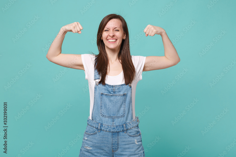 Fototapeta premium Strong smiling young brunette woman girl in casual denim clothes posing isolated on blue turquoise background studio portrait. People lifestyle concept. Mock up copy space. Showing biceps, muscles.