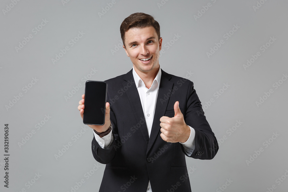 Smiling business man in classic suit shirt isolated on grey background. Achievement career wealth business concept. Mock up copy space. Hold mobile phone with blank empty screen, showing thumb up. Smiling business man in classic suit shirt isolated on grey background. Achievement career wealth business concept. Mock up copy space. Hold mobile phone with blank empty screen, showing thumb up.