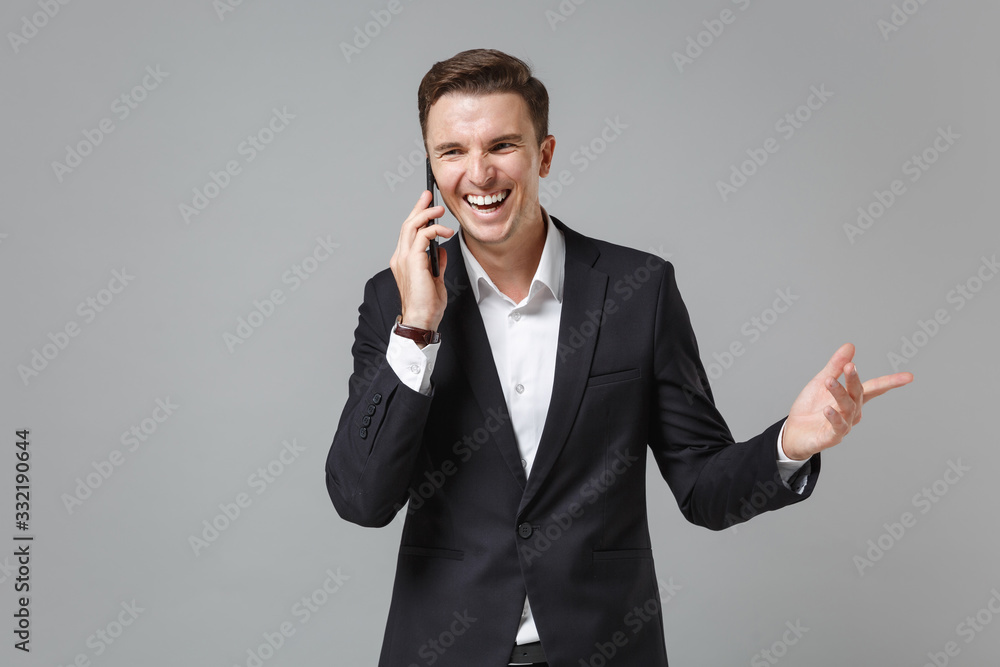 Laughing young business man in classic black suit shirt isolated on grey background in studio. Achievement career wealth business concept. Mock up copy space. Talking on mobile phone spreading hands. Laughing young business man in classic black suit shirt isolated on grey background in studio. Achievement career wealth business concept. Mock up copy space. Talking on mobile phone spreading hands.
