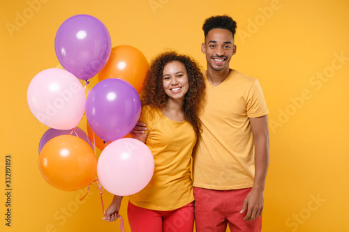 Smiling friends couple african american guy girl in casual clothes isolated on yellow orange background. Birthday holiday party people emotions concept. Celebrating hold colorful air balloons hugging.