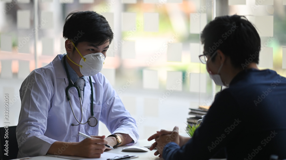Photo of young smart doctor giving an advice to his patient while sitting together at the doctor working desk over orderly examination room as background. Diagnosis/Disease examination concept. Photo of young smart doctor giving an advice to his patient while sitting together at the doctor working desk over orderly examination room as background. Diagnosis/Disease examination concept.
