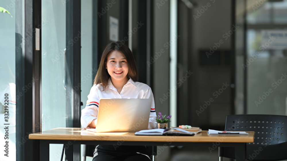 Photo of beautiful girl smiling and sitting in front her computer laptop at the wooden working desk over comfortable living room as background. Photo of beautiful girl smiling and sitting in front her computer laptop at the wooden working desk over comfortable living room as background.