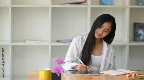 Photo of young beautiful woman in white shirt working with stylus pen and computer tablet while sitting at wooden working table over living room book shelf as background.