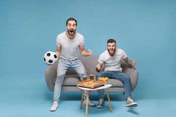 Excited young men guys friends in white t-shirt sit on couch isolated on pastel blue background. Sport leisure concept. Cheer up support favorite team with soccer ball screaming doing winner gesture.