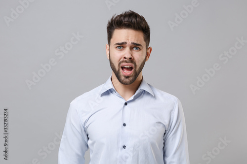 Shocked worried young unshaven business man in light shirt posing isolated on grey wall background studio portrait. Achievement career wealth business concept. Mock up copy space. Looking camera. © ViDi Studio