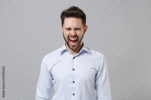 Frustrated crazy young unshaven business man in light shirt posing isolated on grey background in studio. Achievement career wealth business concept. Mock up copy space. Screaming keeping eyes closed. © ViDi Studio