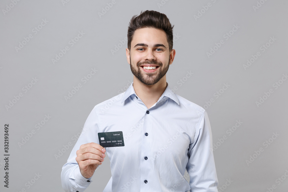 Smiling young unshaven business man in light shirt posing isolated on grey wall background studio portrait. Achievement career wealth business concept. Mock up copy space. Holding credit bank card. Smiling young unshaven business man in light shirt posing isolated on grey wall background studio portrait. Achievement career wealth business concept. Mock up copy space. Holding credit bank card.