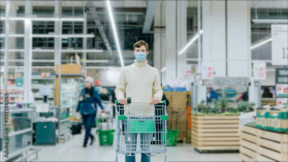 A man in a medical mask stands with a grocery cart in a supermarket, timelapse. Protection from coronavirus, purchase of essential goods. A man in a medical mask stands with a grocery cart in a supermarket, timelapse. Protection from coronavirus, purchase of essential goods.