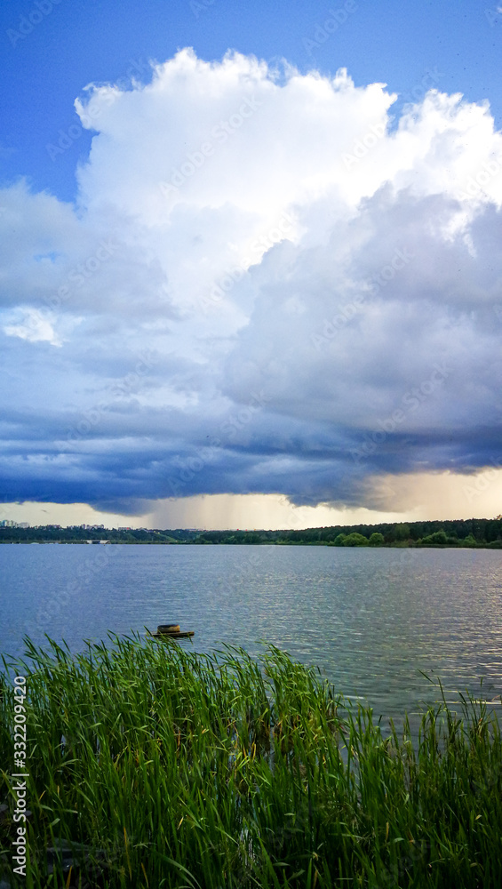 a beautiful blue-white rain cloud over the lake against the blue sky, rain lines are on the horizon, in the foreground of the frame high green grass is waving in the wind