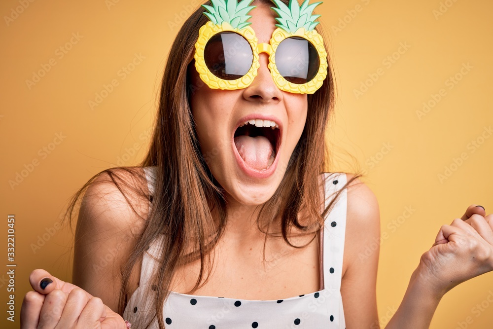 Young beautiful brunette woman wearing pineapple sunglasses over yellow background screaming proud and celebrating victory and success very excited, cheering emotion Young beautiful brunette woman wearing pineapple sunglasses over yellow background screaming proud and celebrating victory and success very excited, cheering emotion