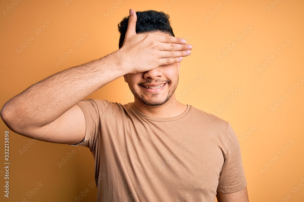 Young handsome man wearing casual t-shirt standing over isolated yellow background smiling and laughing with hand on face covering eyes for surprise. Blind concept. Young handsome man wearing casual t-shirt standing over isolated yellow background smiling and laughing with hand on face covering eyes for surprise. Blind concept.