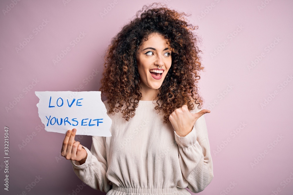 Young beautiful woman with curly hair and piercing holding paper with love yourself message pointing and showing with thumb up to the side with happy face smiling Young beautiful woman with curly hair and piercing holding paper with love yourself message pointing and showing with thumb up to the side with happy face smiling