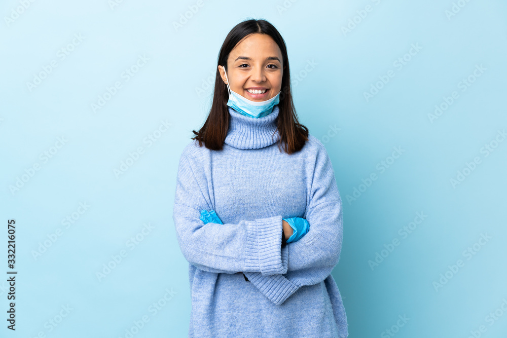 Young brunette mixed race woman protecting from the coronavirus with a mask and gloves over isolated blue background keeping the arms crossed in frontal position Young brunette mixed race woman protecting from the coronavirus with a mask and gloves over isolated blue background keeping the arms crossed in frontal position