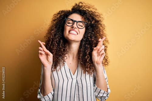 Young beautiful woman with curly hair and piercing wearing striped shirt and glasses gesturing finger crossed smiling with hope and eyes closed. Luck and superstitious concept.