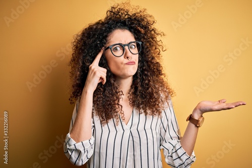 Young beautiful woman with curly hair and piercing wearing striped shirt and glasses confused and annoyed with open palm showing copy space and pointing finger to forehead. Think about it.