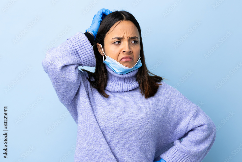 Young brunette mixed race woman protecting from the coronavirus with a mask and gloves over isolated blue background having doubts while scratching head Young brunette mixed race woman protecting from the coronavirus with a mask and gloves over isolated blue background having doubts while scratching head