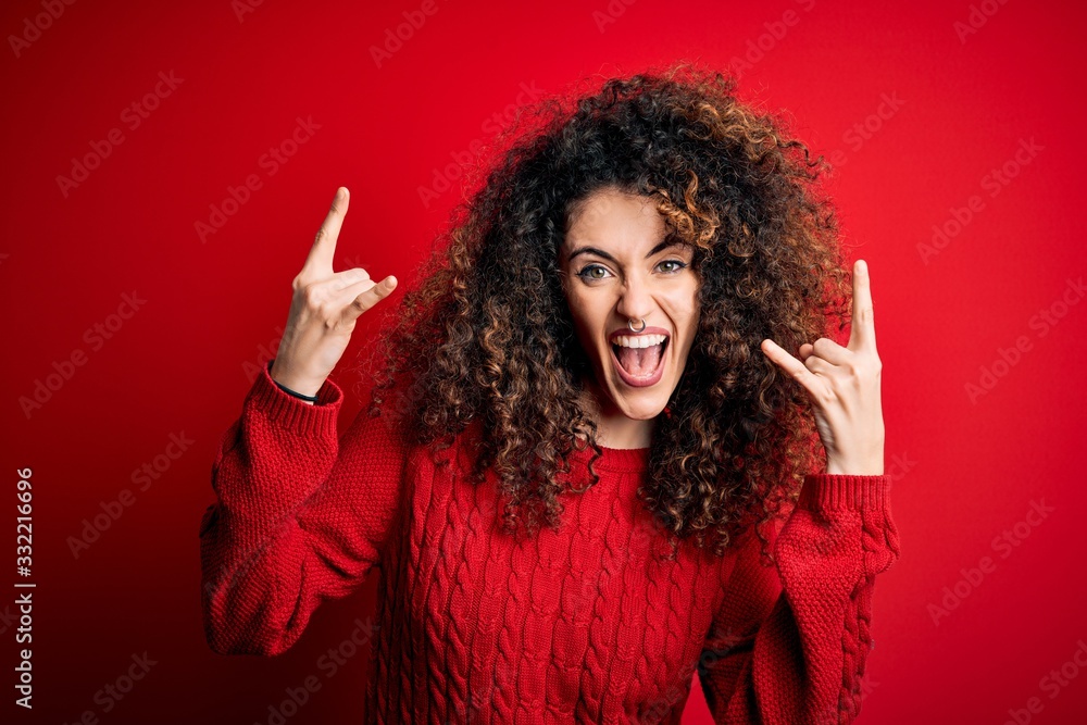 Young beautiful woman with curly hair and piercing wearing casual red sweater shouting with crazy expression doing rock symbol with hands up. Music star. Heavy music concept. Young beautiful woman with curly hair and piercing wearing casual red sweater shouting with crazy expression doing rock symbol with hands up. Music star. Heavy music concept.