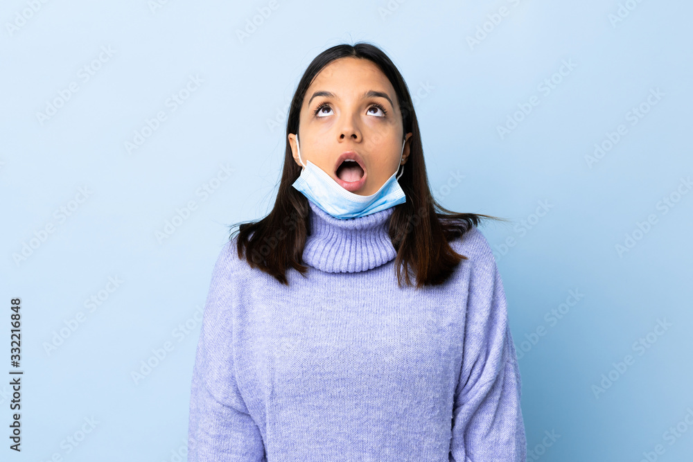 Young brunette mixed race woman protecting from the coronavirus with a mask and gloves over isolated blue background looking up and with surprised expression Young brunette mixed race woman protecting from the coronavirus with a mask and gloves over isolated blue background looking up and with surprised expression
