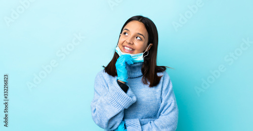Young brunette mixed race woman protecting from the coronavirus with a mask and gloves over isolated blue background looking up while smiling