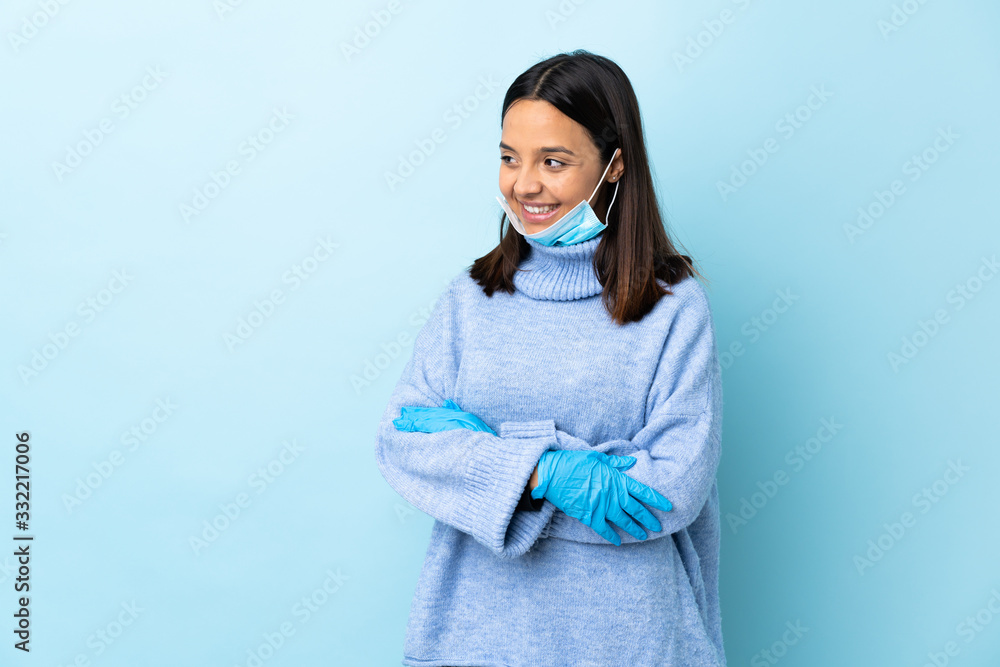 Young brunette mixed race woman protecting from the coronavirus with a mask and gloves over isolated blue background looking to the side and smiling Young brunette mixed race woman protecting from the coronavirus with a mask and gloves over isolated blue background looking to the side and smiling