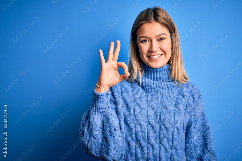 Young beautiful blonde woman wearing winter wool sweater over blue isolated background smiling positive doing ok sign with hand and fingers. Successful expression. Young beautiful blonde woman wearing winter wool sweater over blue isolated background smiling positive doing ok sign with hand and fingers. Successful expression.