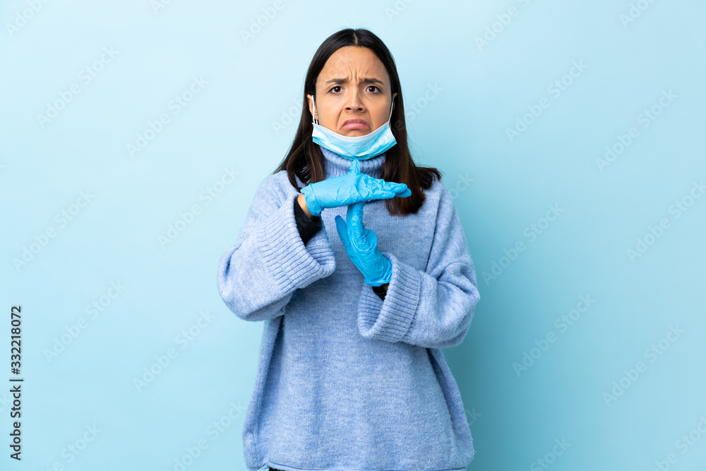 Young brunette mixed race woman protecting from the coronavirus with a mask and gloves over isolated blue background making time out gesture Young brunette mixed race woman protecting from the coronavirus with a mask and gloves over isolated blue background making time out gesture