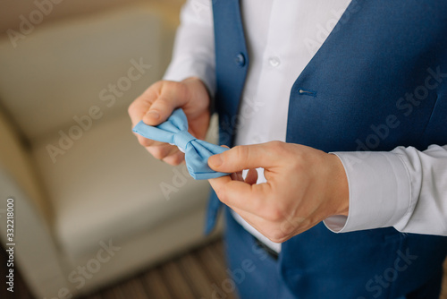 Grooms morning preparation, groom stands near the window and holds bow tie in hands, handsome groom getting dressed and preparing for the wedding. Details. Preparations for wedding. Wedding concept.
