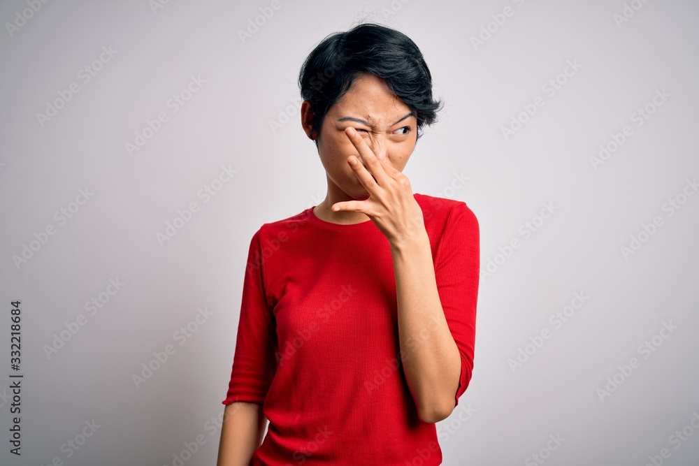Young beautiful asian girl wearing casual red t-shirt standing over isolated white background smelling something stinky and disgusting, intolerable smell, holding breath with fingers on nose Young beautiful asian girl wearing casual red t-shirt standing over isolated white background smelling something stinky and disgusting, intolerable smell, holding breath with fingers on nose