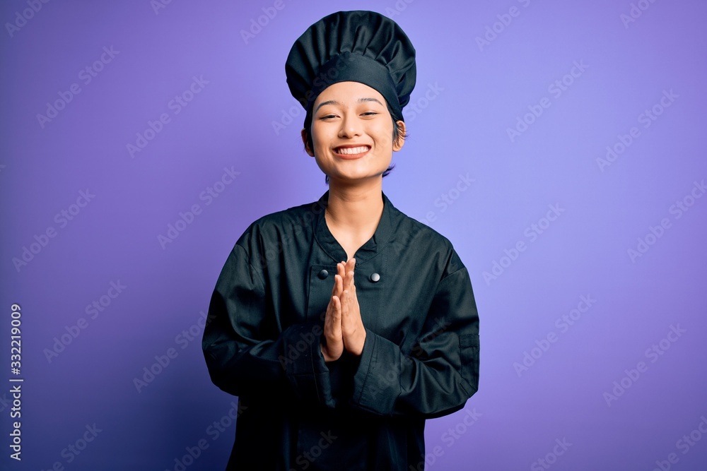 Young beautiful chinese chef woman wearing cooker uniform and hat over purple background praying with hands together asking for forgiveness smiling confident. Young beautiful chinese chef woman wearing cooker uniform and hat over purple background praying with hands together asking for forgiveness smiling confident.