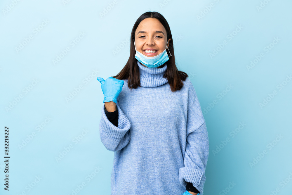Young brunette mixed race woman protecting from the coronavirus with a mask and gloves over isolated blue background pointing to the side to present a product Young brunette mixed race woman protecting from the coronavirus with a mask and gloves over isolated blue background pointing to the side to present a product