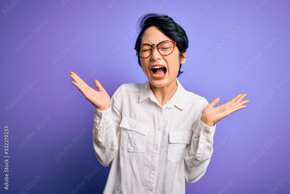 Young beautiful asian girl wearing casual shirt and glasses standing over purple background celebrating mad and crazy for success with arms raised and closed eyes screaming excited. Winner concept Young beautiful asian girl wearing casual shirt and glasses standing over purple background celebrating mad and crazy for success with arms raised and closed eyes screaming excited. Winner concept