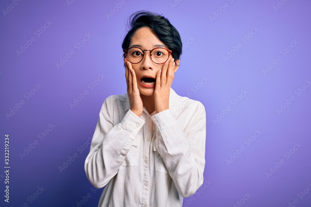 Young beautiful asian girl wearing casual shirt and glasses standing over purple background afraid and shocked, surprise and amazed expression with hands on face Young beautiful asian girl wearing casual shirt and glasses standing over purple background afraid and shocked, surprise and amazed expression with hands on face