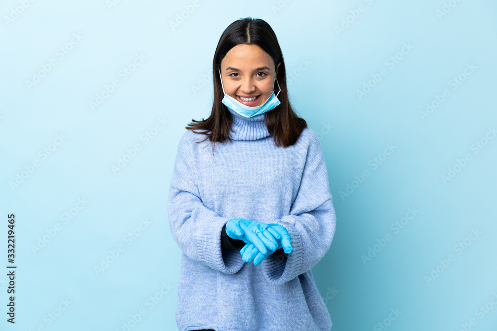 Young brunette mixed race woman protecting from the coronavirus with a mask and gloves over isolated blue background holding copyspace imaginary on the palm to insert an ad Young brunette mixed race woman protecting from the coronavirus with a mask and gloves over isolated blue background holding copyspace imaginary on the palm to insert an ad