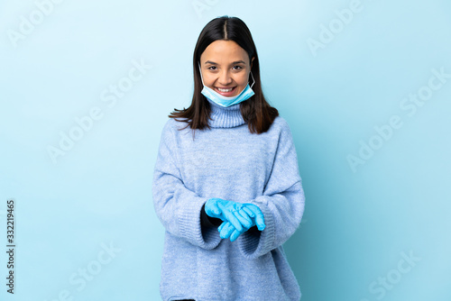 Young brunette mixed race woman protecting from the coronavirus with a mask and gloves over isolated blue background holding copyspace imaginary on the palm to insert an ad