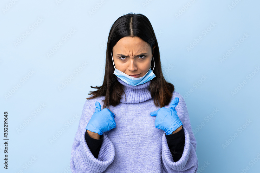 Young brunette mixed race woman protecting from the coronavirus with a mask and gloves over isolated blue background pointing to oneself Young brunette mixed race woman protecting from the coronavirus with a mask and gloves over isolated blue background pointing to oneself