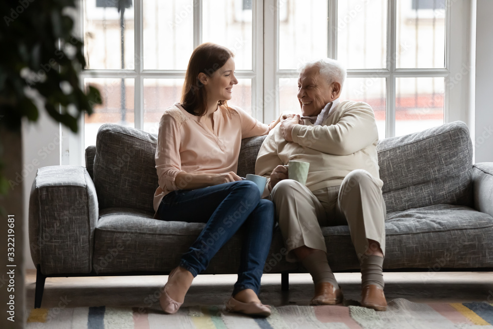 In cozy living room caring adult daughter drink tea with old dad family enjoy talk sit on comfy couch. Caregiver and patient care, attention, love, strong connection with older relative person concept