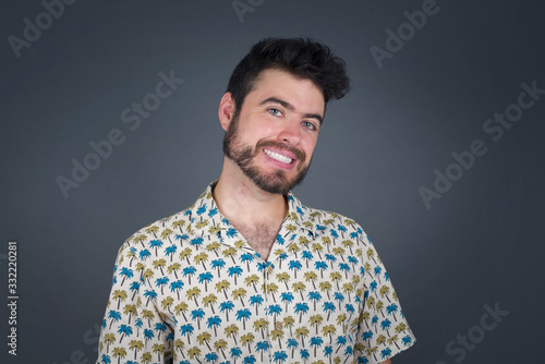 Shy blond boy smiling looking to the camera. Caucasian charming young man sanding against gray studio background feeling shy.