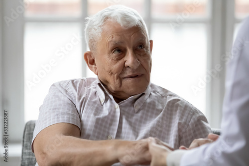 Woman nurse hold hand of elderly grey-haired man showing care and bond close up, doctor in white coat talk with aged patient sitting together indoor, concept of support, caregiving and nursing service