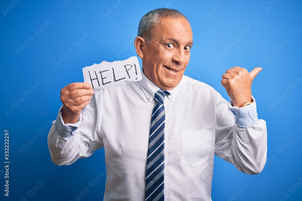 Senior handsome businessman holding paper help message over isolated blue background pointing and showing with thumb up to the side with happy face smiling Senior handsome businessman holding paper help message over isolated blue background pointing and showing with thumb up to the side with happy face smiling