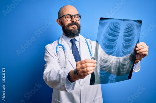 Handsome bald doctor man with beard wearing stethoscope holding chest xray with a happy face standing and smiling with a confident smile showing teeth