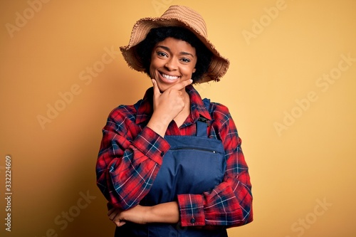 Young African American afro farmer woman with curly hair wearing apron and hat looking confident at the camera smiling with crossed arms and hand raised on chin. Thinking positive.