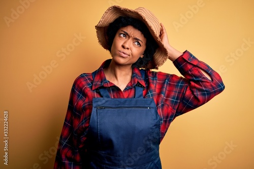 Young African American afro farmer woman with curly hair wearing apron and hat confuse and wonder about question. Uncertain with doubt, thinking with hand on head. Pensive concept.