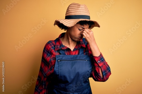 Young African American afro farmer woman with curly hair wearing apron and hat tired rubbing nose and eyes feeling fatigue and headache. Stress and frustration concept.