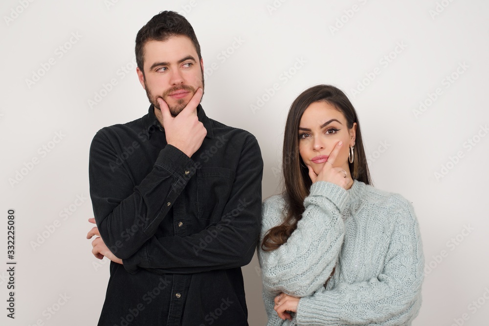 Isolated portrait of stylish young European woman with hand under chin and looking sideways with doubtful and skeptical expression, worry and doubt. Standing indoors over gray background. Isolated portrait of stylish young European woman with hand under chin and looking sideways with doubtful and skeptical expression, worry and doubt. Standing indoors over gray background.