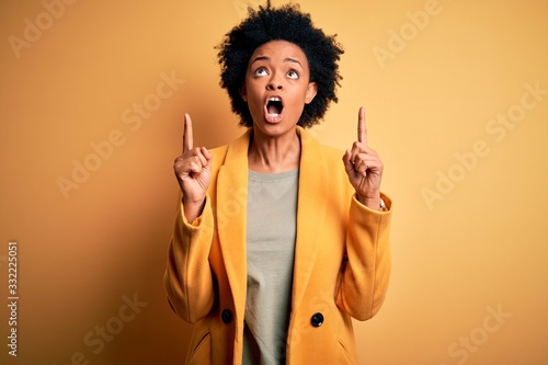 Young beautiful African American afro businesswoman with curly hair wearing yellow jacket amazed and surprised looking up and pointing with fingers and raised arms.